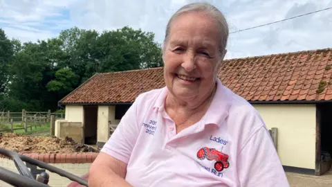 Jenny Kirk/BBC Mrs Chapman is smiling at the camera and wearing her Ladies' Road Run polo shirt. She is sitting at the wheel of a tractor and there are farm buildings and a field behind her.