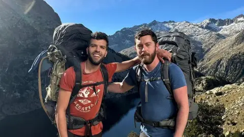 File photo of Aziz Ziriat (left) and Sam Harris in shorts and T-shirts wearing large hiking backpacks and stood on top of a hilltop overlooking a gorge with their arms on each others' shoulders.