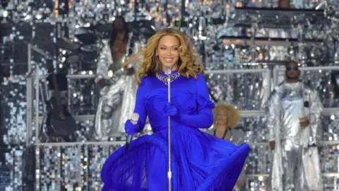 Getty Images Image of Beyoncé wearing a blue dress as she sings at Tottenham Stadium in 2023 with a silver background and band behind her