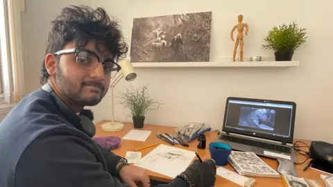 Aaban sits at a desk. On the wall is a photograph in black and white taken by the photographer James Ravillious of a woman herding sheep. On the desk he has ink, paper and sketches for his graphic novel, Widening Gyre.