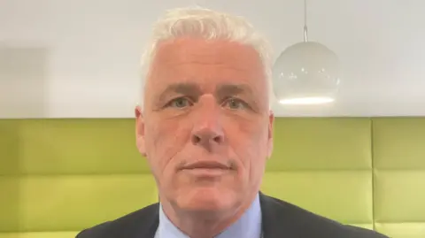 Fearghal McKinney, from the BHF, is wearing a achromatic  suit   with a bluish  garment  and a reddish  patterned tie. He's lasting  down  brushed  greenish  walls.