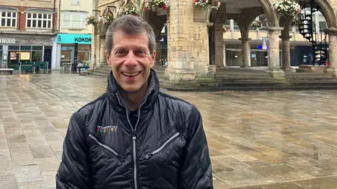 Shariqua Ahmed /BBC Pep wearing a black jacket smiling for the camera standing in front of a historic guildhall