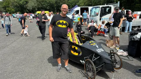 Stuart Hinchcliffe stood in a car park next to a homemade Batman-inspired kart. He is wearing a black Batman T-shirt, black shorts and grey trainers. He has a bald head and is wearing black-rimmed glasses. Groups of people are walking, standing or sitting behind him. There are also other parked vehicles behind. 