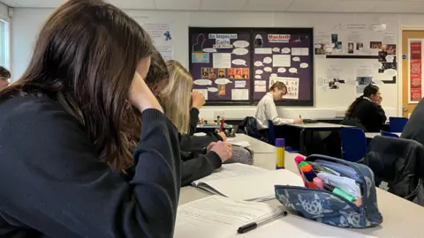 BBC Hay una fila de estudiantes en un salón de clases en saltadores de la Marina con la cabeza hacia abajo mirando sus cuadernos. Una caja de lápiz está sobre la mesa. 