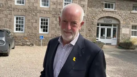 Steve Luce pictured in front of the granite buildings of the environment department. He is wearing a navy blazer and white and purple pinstripe shirt. He has short white hair and stubble. 