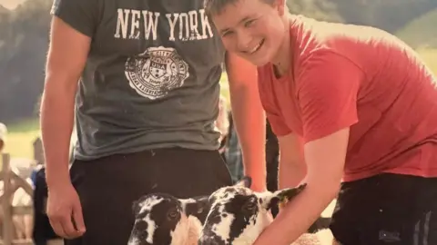 Jordan Burrow An old photo of Jordan when he was younger. He is crouching over two sheep and smiling into the camera. He is wearing a red t-shirt and standing in a field. Another person is standing beside him in a t-shirt that reads 'New York'.