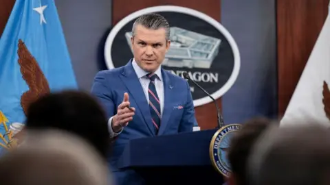 Pete Hegseth stands at a podium wearing a blue suit and a blue striped tie while point his right hand towrads reporters