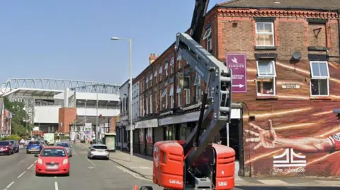 Liverpool FC's stadium is in the background as cars drive the street. On the right is a wall with part of a mural showing the hand of former Liverpool player Steven Gerard.