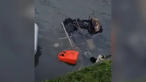 A pile of burnt debris, including an orange jerry can and what is left of a gutted canal boat, on a canal, near the grass bank.