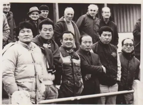 Phil Monckton A black and white photo of several Korean men standing in a rugby stand in thick jackets.