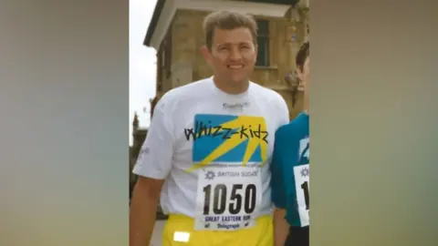 Supplied A runner wearing a white ‘Whizz‑Kidz’ event T‑shirt with a race bib number 1050 and bright yellow shorts stands outdoors in front of a stone building.