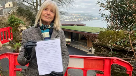 Marina Pusey, from Warfleet Road Warriors, holding the petition paper. She has short blonde hair and is wearing a blue and white scarf, a grey jacket and black gloves. She is smiling at the camera.