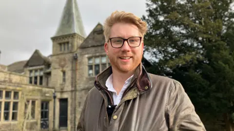 BBC/Mark Ansell Jake Dinsdale is standing in front of his hotel Simonstone Hall near Hawes. He has short fair hair and a short beard and is wearing glasses.