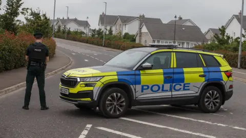 Pacemaker Police officer facing away from the camera standing next to a police car parked in the middle of a road with some houses in the background