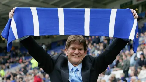 Sir John Madejski, holding a blue and white scarf above his head, celebrating Reading's promotion to the Premier League against Leicester in 2006.