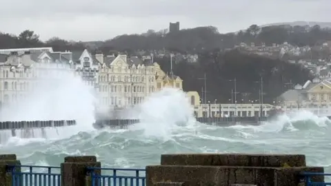 Waves crashing against railings on Douglas Promenade. The Victorian seafront buildings line the promenade behind.