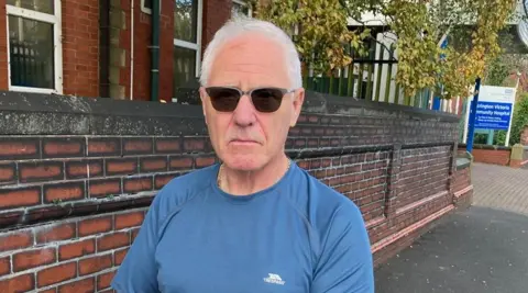 Steve Richards has grey hair and is wearing sunglasses and a light blue T-shirt. He is standing in front of a brick wall at the entrance to Accrington Victoria Community Hospital.