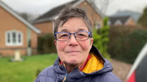 A woman with short dark hair, wearing glasses and a blue coat looks into the camera with homes behind her.