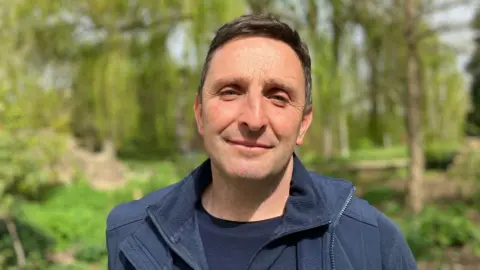 Joe, a man with short brown hair, wearing a blue T-shirt and a blue jacket, smiling for camera.