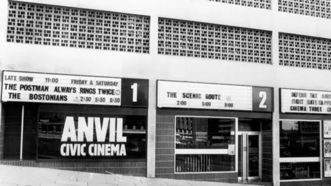 Estate by Dave Godin A black and white image of a city street showing three commercial units with Anvil Civic Cinema in large letters on one unit's window. All three units have billboards above the main windows displaying movie titles and screening times. 