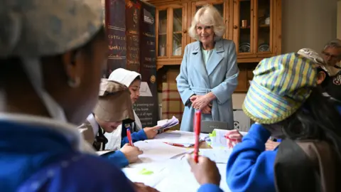 PA Media Queen Camilla wearing a pale green coat stands in front of a table where school children in Victorian-era costumes sit writing.