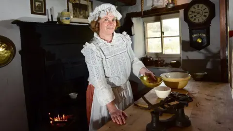 Anthony Chappel-Ross A woman wearing traditional Victorian costume - white floaty shirt, red long skirt and a white bonnet hat - measures out ingredients, in a kitchen.