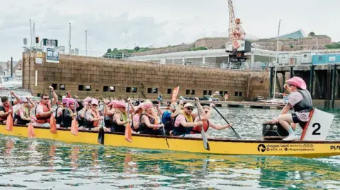 Jersey Hospice Care A photo that shows a number of people in a long yellow boat, with one man at the front of the boat facing the other people. He is wearing a pink hat. Some of the other people are also wearing pink hats or wigs. They are all holding oars that are out of the water.