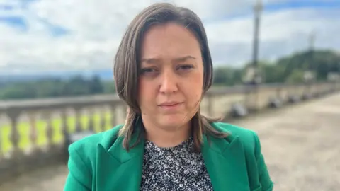 A woman is standing outside. She has shoulder-length brown hair. She's wearing a green jacket and a blue and white top.