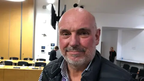 Vikki Irwin/BBC A picture of Dave Taylor. He is facing the camera and smiling. He is standing in a council room with tables and chairs behind him. He is bald and has a grey beard. He is wearing a blue gilet jacket over the top of a striped blue and white shirt.