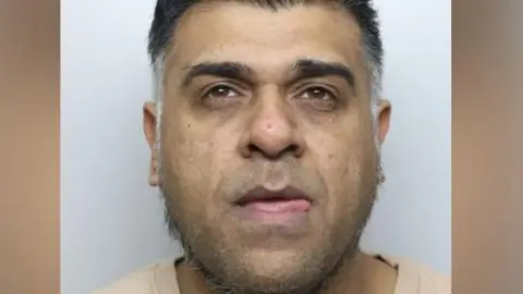 A police mugshot of a man in his 40s. He has greying hair and is wearing a beige T-shirt. He is staring ahead above the camera.