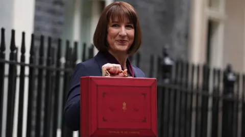 PA The chancellor, Rachel Reeves, standing in Downing Street holding the Chancellor of the Exchequer red box. She is wearing a dark suit with a red shirt. 