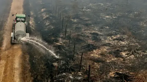 A drone view shows a farmer using his tractor to spray burnt fields and forests during a wildfire burning in the High Fens