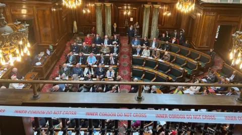 Aisha Iqbal/BBC A bird's eye view of a full council chamber. Men and women sit in a series of horseshoe-shaped wooden benches facing a table at one end. The wood-paneled room is lit by a number of chandeliers hanging from the ceiling.