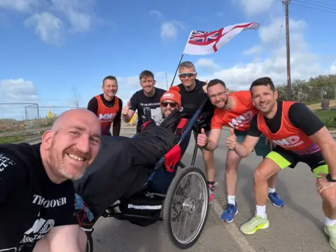 A group of men smile in a selfie wearing orange t-shirts surrounding Jon in a wheelchair.