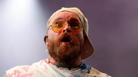 Teddy Swims with a light brown beard wearing a beige baseball cap sideways and sunglasses, pearl necklace and a white tye-dye t-shirt. He is on stage performing and looks surprised with his eyes wide and mouth open facing towards the crowd at Radio 1's Big Weekend festival.