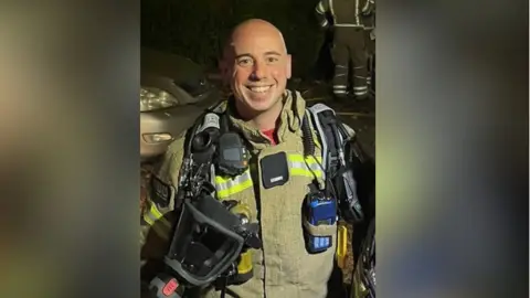 Firefighter Martyn Sadler stands in his uniform looking at the camera. He has firefighting equipment hanging off his uniform. He is smiling.