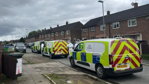 Davinia Ramos/BBC Three police vans are parked on a residential street. Two forensic officers in full white suits and hair nets are walking alongside the vans.