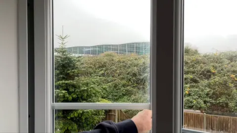 A view out of a window from inside a house. An arm holds the window open. Behind, you can see a garden with trees and bushes, and the new warehouse building poking over the top.