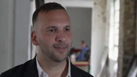Zack Polanski being interviewed for TV. He is wearing a dark jacket and a white open-necked shirt. There is an blurred background image with a window to one side. 