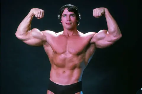 Getty Images Arnold Schwarzenegger is his younger body-building days, he is wearing black trunks and is flexing his muscles.