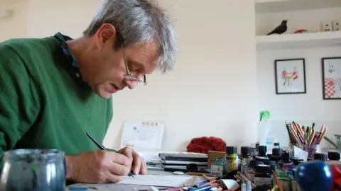 Axel Scheffler pinta en su estudio. Lleva gafas y sostiene un bolígrafo sobre su escritorio, donde está rodeado de botes de bolígrafos, tinta y lápices.