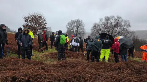 Richard Clarke A picture of a group of around 30 people gathered on a mountain side with grey sky surrounding them, appearing sodden with rain. Some have dogs on short leads and others have walking sticks. 
Around three people can be seen holding umbrellas while everyone wears raincoats and has their hood up. 