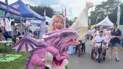 A participant is holding a bright pink and purple dragon. They have blonde hair and are dressed in medieval costume.
