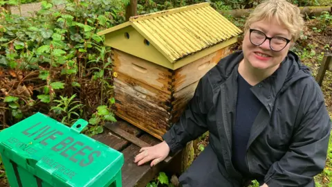 A Woman sits next to a beehive and a green box labelled 'Live Bees'.