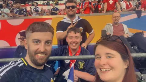 Stephen Reside A smiling Stephen (centre) sits in the stand for a selfie style pic taken by a fellow supporter. He is wearing a Scotland shirt. A man and woman, both smiling, are in the foreground. A  smiling man is standing behind Stephen and making a v-sign behind his head.