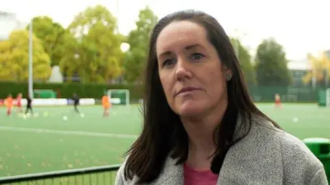 Ria Burrage-Male at the side of a hockey pitch. She has shoulder length straight black hair and is wearing a grey sweater over a pink t-shirt