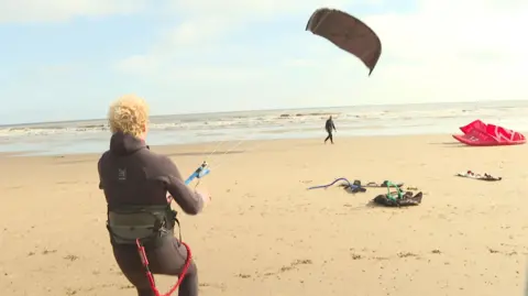 A kitesurfer in a black wetsuit leans back as he controls a black kite, which is rising from the beach, with the sea in the background