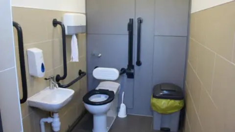 Inside of a refurbished accessible toilet cubicle. The walls feature beige tiles and the back wall is painted grey. The toilet is on the left and is surrounded by handles. The sink is also on the right. There is a bin with a yellow bin bag on the right. 