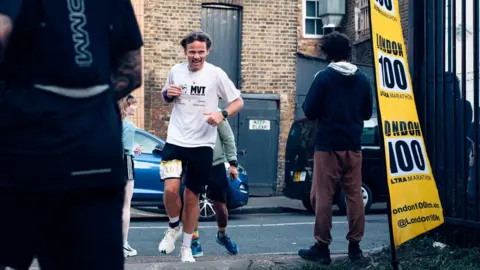 Derrick Wess Andy wears a white top and black shorts. He is running along a street. On the right is a yellow banner, which says London 100 Ultra Marathon.