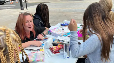 John Devine/BBC Katy Shroff and three anonymous teenage girls at a table in Broad Street, March. The table is covered in colouring pens and pencils, plus a couple of packets of crisps. Katy is on the far left, has crouched down at the table and looking towards the viewer and smiling. She has reddish shoulder-length hair, with her glasses perched on her head and is wearing a black top. The three girls all have long hair and have turned their backs to the viewer. 
